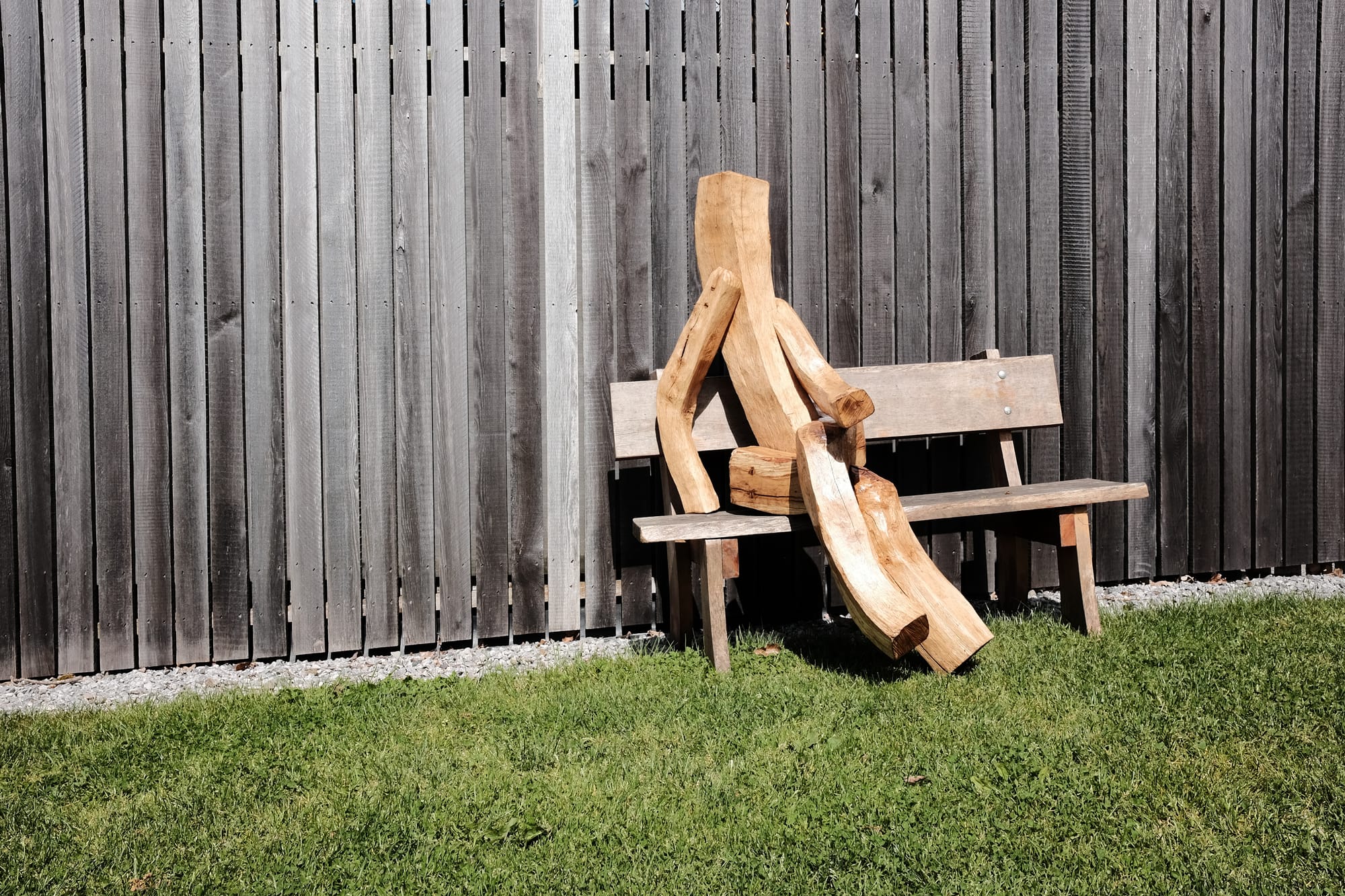 A light brown, angular wooden sculpture of a human figure sits on a brown wooden bench against the grey wooden slats of a barn