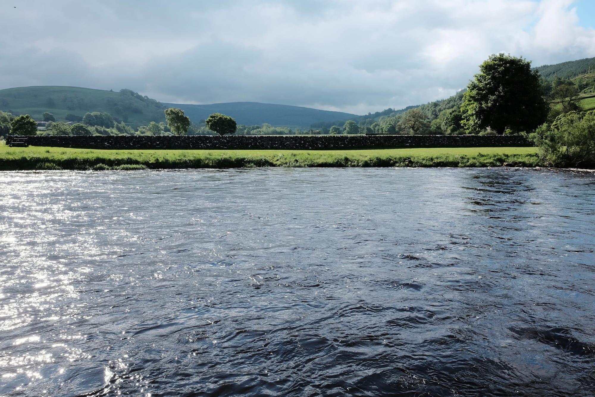 The view of the River Wharfe from Burnsall, water lapping at grassy banks with a stone wall and high ground in the distance.
