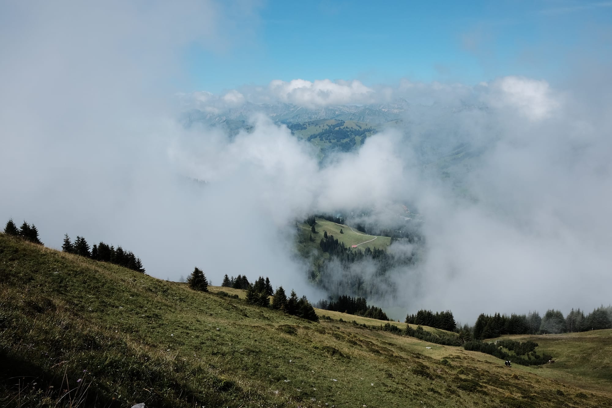 The grassy slopes of a mountain with a break in the low cloud revealing a red-roofed building in a meadow below.