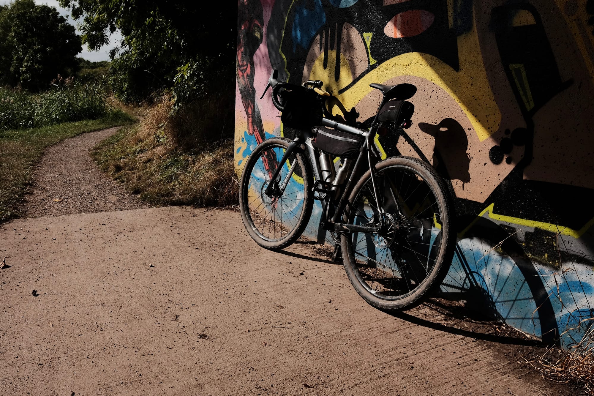 A grey bike leans against a concrete wall covered in multi-coloured graffiti, beyond which lies a narrow stone trail.