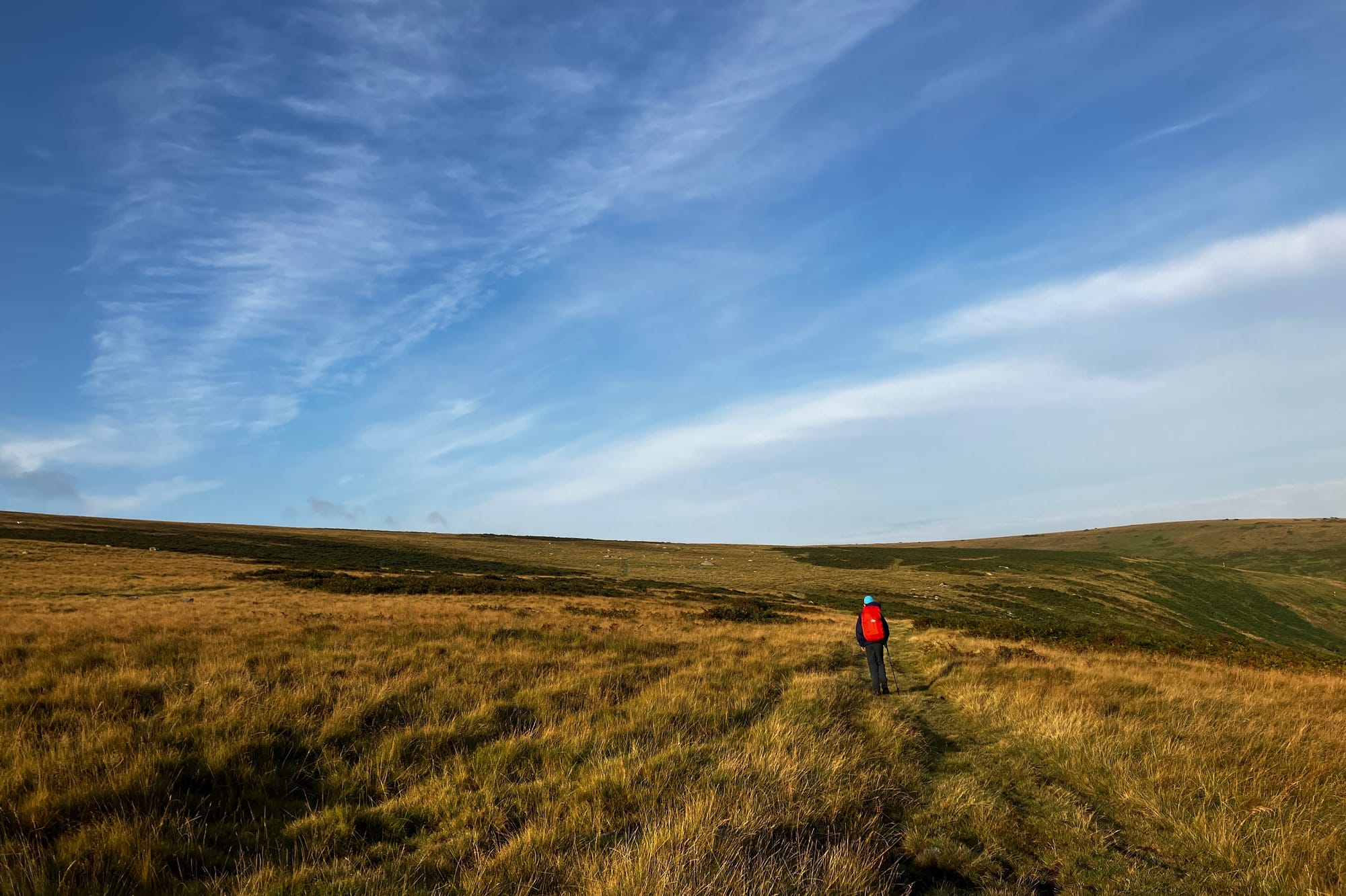 A child with e red rucksack walks away along a moorland path, a large hill visible in the distance against blue sky.