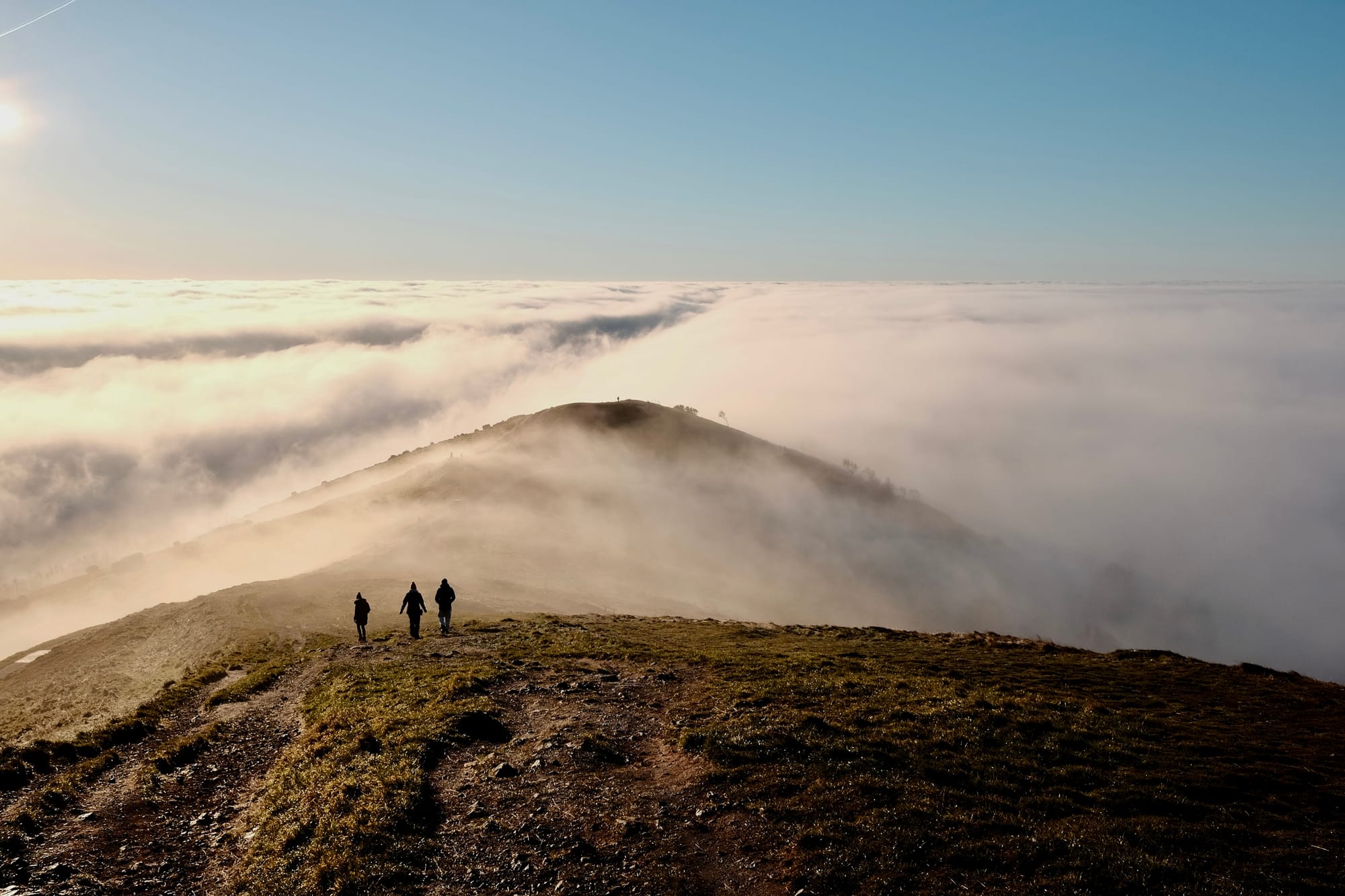 Three figures walk down a hilltop ridgeline with low cloud stretching to the horizon.