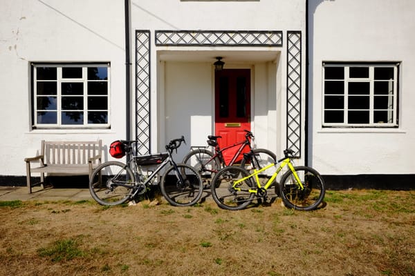 Three bikes – one grey, one red and black, and one bright yellow, lean against the porch of a white house with a red door.